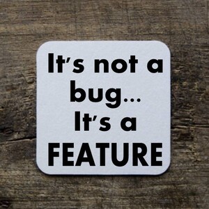 May include: White square coaster with black text that reads "It's not a bug... It's a FEATURE".
