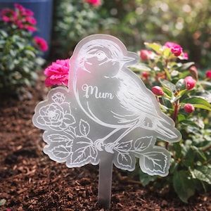 Puede incluir: Un marcador de jardín de acrílico transparente con un diseño de pájaro y flores, y la palabra "Mum" en escritura cursiva. El marcador está colocado en un jardín con flores rosas y tierra marrón. El marcador es de metal.