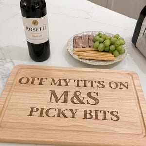 Può includere: Un tagliere in legno con il testo inciso "OFF MY TITS ON M&S PICKY BITS". Una bottiglia di vino Rosetti Merlot, un piatto di uva, grissini e salumi sono anch'essi presenti.