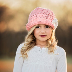 May include: A young girl with blonde hair wearing a pink crocheted hat with a white flower accent and a white lace-trimmed shirt.