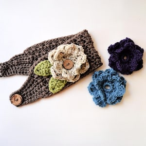 May include: A dark brown crocheted headband featuring a cream-colored crocheted flower with two light green leaves and a wooden button center. The headband has a wooden button closure. Two additional crocheted flowers in dark purple and blue are included.