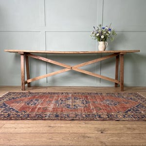 May include: A long, narrow wooden console table with a cross-brace base design. A white ceramic pitcher filled with a bouquet of wildflowers sits on the table. A rectangular area rug with a red, blue, and tan patterned design lies on the floor.
