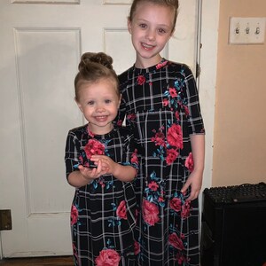 May include: Two young girls wearing black and white plaid dresses with floral accents. The dresses have a long, flowing silhouette and feature a red, pink, and blue floral pattern.