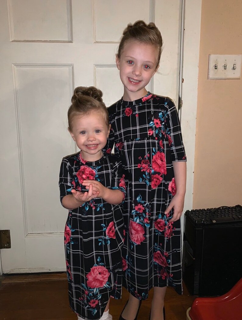 May include: Two young girls wearing black and white plaid dresses with floral accents. The dresses have a long, flowing silhouette and feature a red, pink, and blue floral pattern.