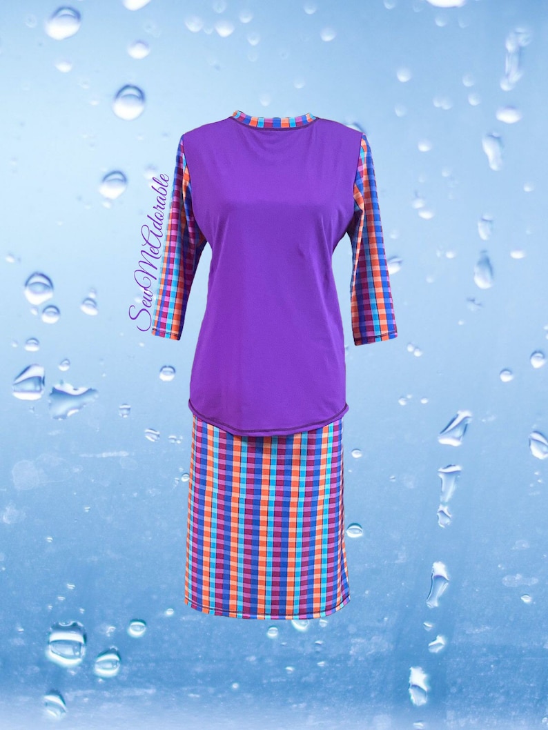 May include: A two-piece outfit consisting of a solid purple long-sleeve top and a matching skirt. The sleeves and skirt feature a vibrant plaid pattern in orange, blue, and purple. The text SewMeAdorable is printed on the left side of the top.