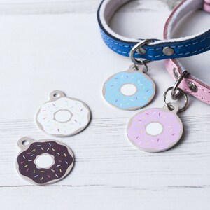 May include: Four dog tags with different colored donut designs. The donuts are white with pink frosting and sprinkles, white with blue frosting and sprinkles, white with chocolate frosting and sprinkles, and pink with white frosting and sprinkles.