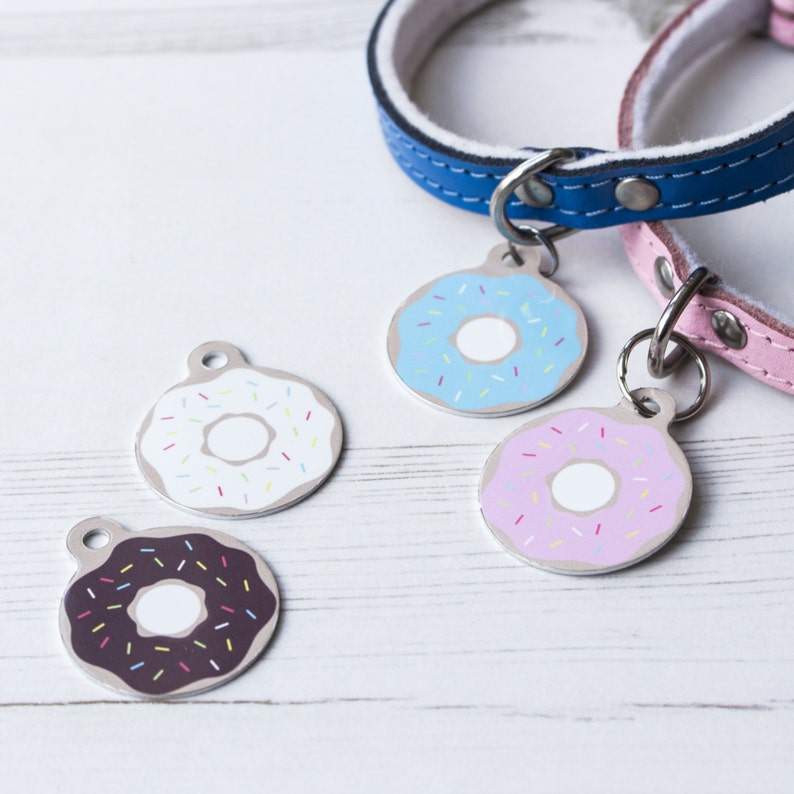 May include: Four dog tags with different colored donut designs. The donuts are white with pink frosting and sprinkles, white with blue frosting and sprinkles, white with chocolate frosting and sprinkles, and pink with white frosting and sprinkles.