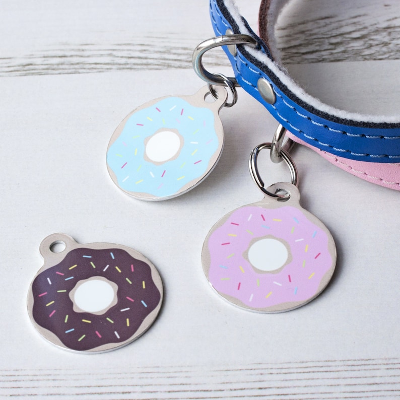 May include: Three dog tags with donut designs. The donuts are blue with white frosting and sprinkles, pink with white frosting and sprinkles, and brown with white frosting and sprinkles.