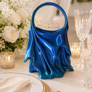May include: A vibrant blue handbag featuring a circular top handle and a draped, fabric-like sculptural design. The surface has a glossy, metallic finish that transitions into a darker teal hue at the base, creating a fluid, rippled appearance.