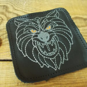 Könnte beinhalten: Schwarzer Lederuntersetzer mit einem weißen gestickten Wolfskopf mit orangefarbenen Augen. Der Wolf knurrt mit sichtbaren Zähnen.
