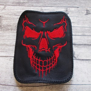 Könnte beinhalten: Ein schwarzer Leder-Patch mit einem roten gestickten Totenkopf-Design. Der Totenkopf hat rote Augen und einen detaillierten Mund.