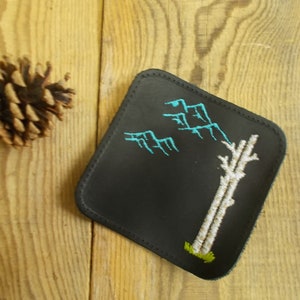 May include: Black leather coaster with a white tree and blue mountains embroidered on it.