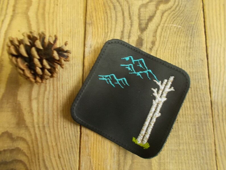 May include: Black leather coaster with a white tree and blue mountains embroidered on it.