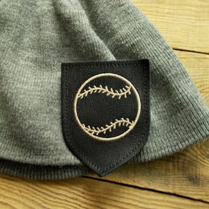 May include: A gray beanie with a black patch featuring a baseball design. The baseball is embroidered in a cream color, with the stitching details clearly visible. The beanie is made of a soft, knitted material.