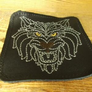 Könnte beinhalten: Ein schwarzer Leder-Patch mit einem weißen gestickten Bild eines knurrenden Wolfskopfes mit gelben Augen.