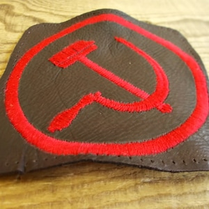 May include: A red embroidered hammer and sickle patch on a brown leather background. The patch is surrounded by a red circle.