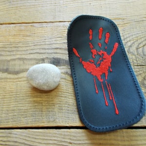 May include: A black leather eyeglass case with a red handprint design. The handprint is embroidered with dripping blood details. A small, smooth, gray stone sits next to the case on a wooden surface.
