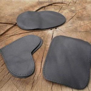 Könnte beinhalten: Drei graue Lederartikel sind auf einer Holzoberfläche angeordnet. Die Artikel umfassen eine abgerundete Form, eine Herzform und eine quadratische Form. Jeder Artikel hat einen genähten Rand, was auf handwerkliche Arbeit hindeutet.