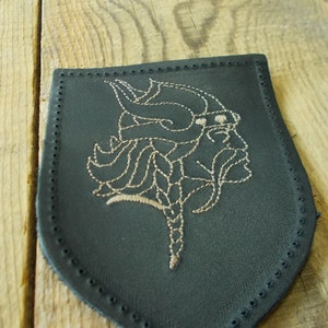 Może przedstawiać: Czarna skórzana łatka z wyszytym wzorem wikinga wojownika w rogacym hełmie. Wzór wykonany jest jasnobrązową nicią.