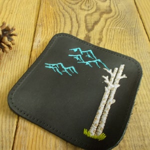 May include: A black leather coaster with a white embroidered tree and blue mountains. The coaster has a stitched edge.