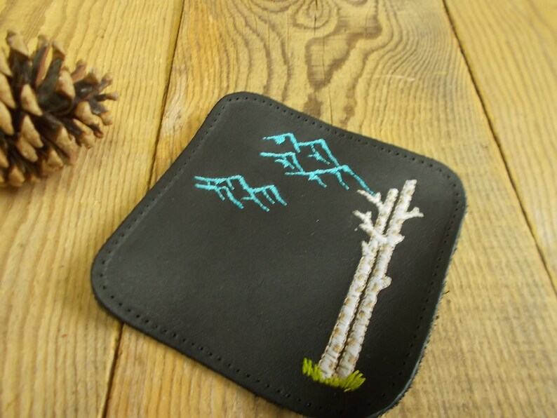 May include: A black leather coaster with a white embroidered tree and blue mountains. The coaster has a stitched edge.