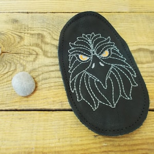 以下が含まれることがあります： 黒い革のパッチに刺繍された鷲の頭のデザイン。鷲は白いステッチとオレンジ色の目をしています。