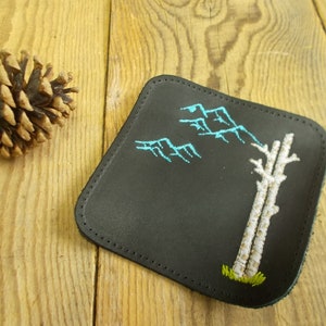 May include: Black leather coaster with white and blue embroidery of a tree and mountains.