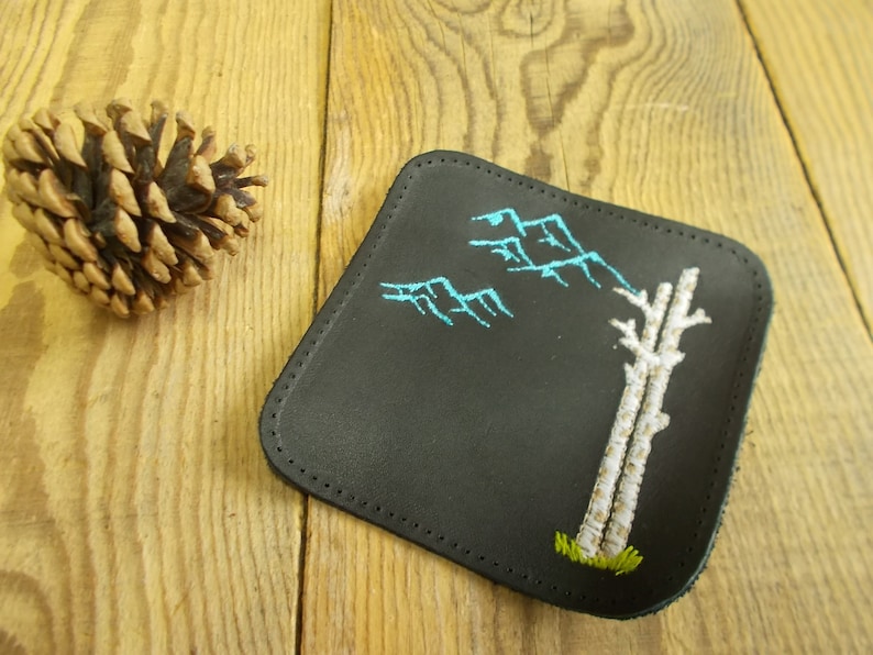 May include: Black leather coaster with white and blue embroidery of a tree and mountains.