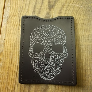 Könnte beinhalten: Ein schwarzer Lederkartenhalter mit einem weißen gestickten Totenkopfdesign. Der Totenkopf ist mit komplizierten Wirbeln und Mustern verziert.