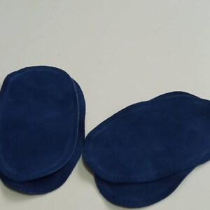 May include: Two pairs of blue suede elbow patches. Each patch is oval shaped and has a stitched edge.