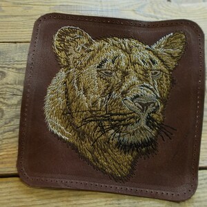 Puede incluir: Parche bordado con la cabeza de un león sobre un fondo de cuero marrón. El pelo del león es una mezcla de hilo dorado y marrón.