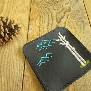May include: Black leather coaster with white embroidered trees and blue embroidered mountains.