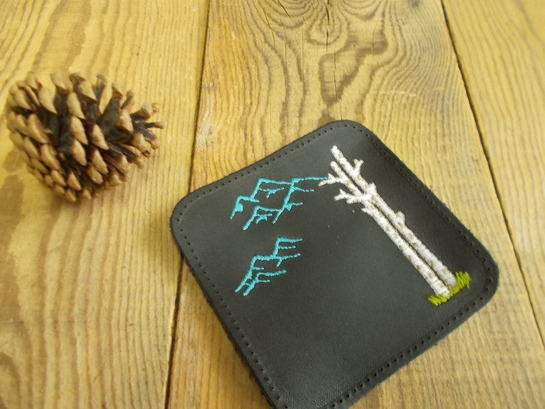 May include: Black leather coaster with white embroidered trees and blue embroidered mountains.