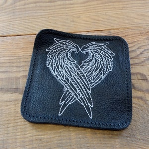 May include: Black leather coaster with white embroidered angel wings forming a heart shape.