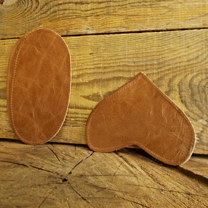 Puede incluir: Dos parches de cuero marrón, uno de forma ovalada y otro en forma de corazón, sobre una superficie de madera.
