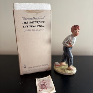 May include: A figurine of a young boy in blue pants and a striped shirt, part of the "Norman Rockwell" The Saturday Evening Post Cover Collection. The figurine is on a small base, with a miniature magazine nearby. The packaging is also visible.