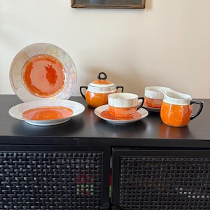 May include: A vintage tea set with orange and white geometric designs. The set includes a large plate, a smaller plate, a sugar bowl, two cups, two saucers, and a creamer. The plates have a pearlescent rim. The set is displayed on a black cabinet.