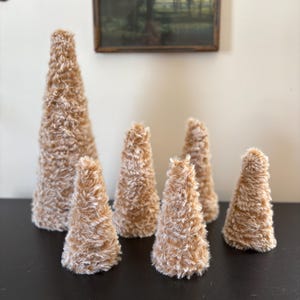 May include: A collection of six decorative, cone-shaped objects with a fuzzy, light brown texture. The cones vary in size, with the tallest one standing prominently in the background. They are arranged on a dark surface, suggesting a display or decorative arrangement.