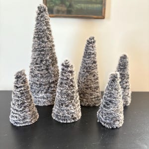 May include: A collection of six decorative cone-shaped trees. The trees are covered in a soft, textured, gray and white faux fur. The cones vary in size, creating a visually interesting display. The trees are arranged on a black surface.