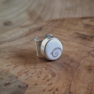 Puede incluir: Un anillo de plata con una incrustación de concha blanca. La concha tiene un patrón en espiral.