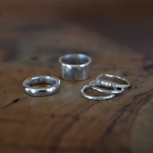 Könnte beinhalten: Vier silberne Ringe auf einer Holzoberfläche. Ein Ring ist breiter als die anderen und hat eine glatte, abgerundete Oberfläche. Die anderen drei Ringe sind dünner und haben eine strukturierte Oberfläche.