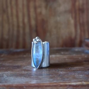 Peut inclure: Une bague en argent avec une pierre de lune bleue sertie dans une bande d'argent. La bague est posée sur une surface en bois.