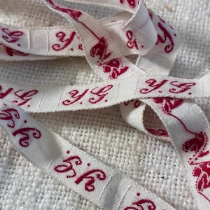 May include: A white fabric ribbon featuring a repeating pattern of the letters y.g. printed in a dark pink script font. The ribbon is folded and layered, showcasing the continuous text design along its length against a textured white background.