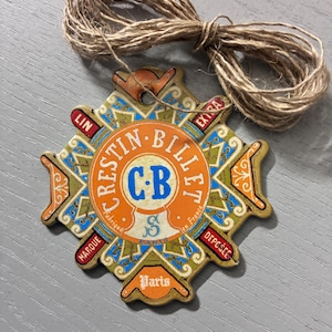 May include: A decorative star-shaped cardboard tag with a vintage-style label featuring the text Crestin-Billet, C.B, Paris, Lin, Extra, Marque, and Deposee. The design uses orange, blue, and gold colours, and includes a loop of brown twine for hanging.