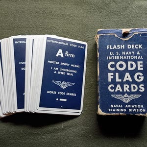 May include: Vintage flash cards for naval aviation training. The deck of cards features blue cards with white text and illustrations. The box reads "Flash Deck U.S. Navy & International Code Flag Cards". The cards are for learning international code flags.