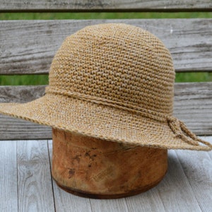 May include: A tan crochet hat with a wide brim and a decorative band around the crown. The hat is displayed on a wooden form, showcasing its shape and texture. The brim is slightly curved, and the hat has a casual, summery style.