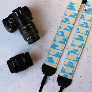 May include: A black DSLR camera with a lens and a separate lens next to a camera strap. The camera strap is cream-coloured with a blue mountain pattern and black accents. The strap is designed for a camera.