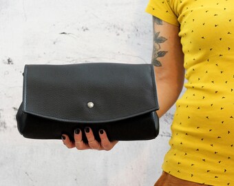 May include: A black leather clutch with a flap closure and a silver button. The clutch is being held by a person's hand.