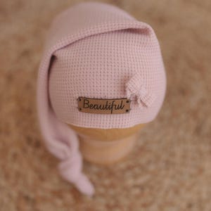 May include: A light pink baby bonnet with a textured waffle knit design. The bonnet has a long, draped tail and a small bow detail. A small brown tag with the word "Beautiful" is attached to the front.