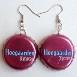 Puede incluir: Un par de pendientes hechos de tapas de botellas rosas con las palabras "Hoegaarden Rosée" impresas en ellas. Los pendientes están unidos a ganchos plateados.
